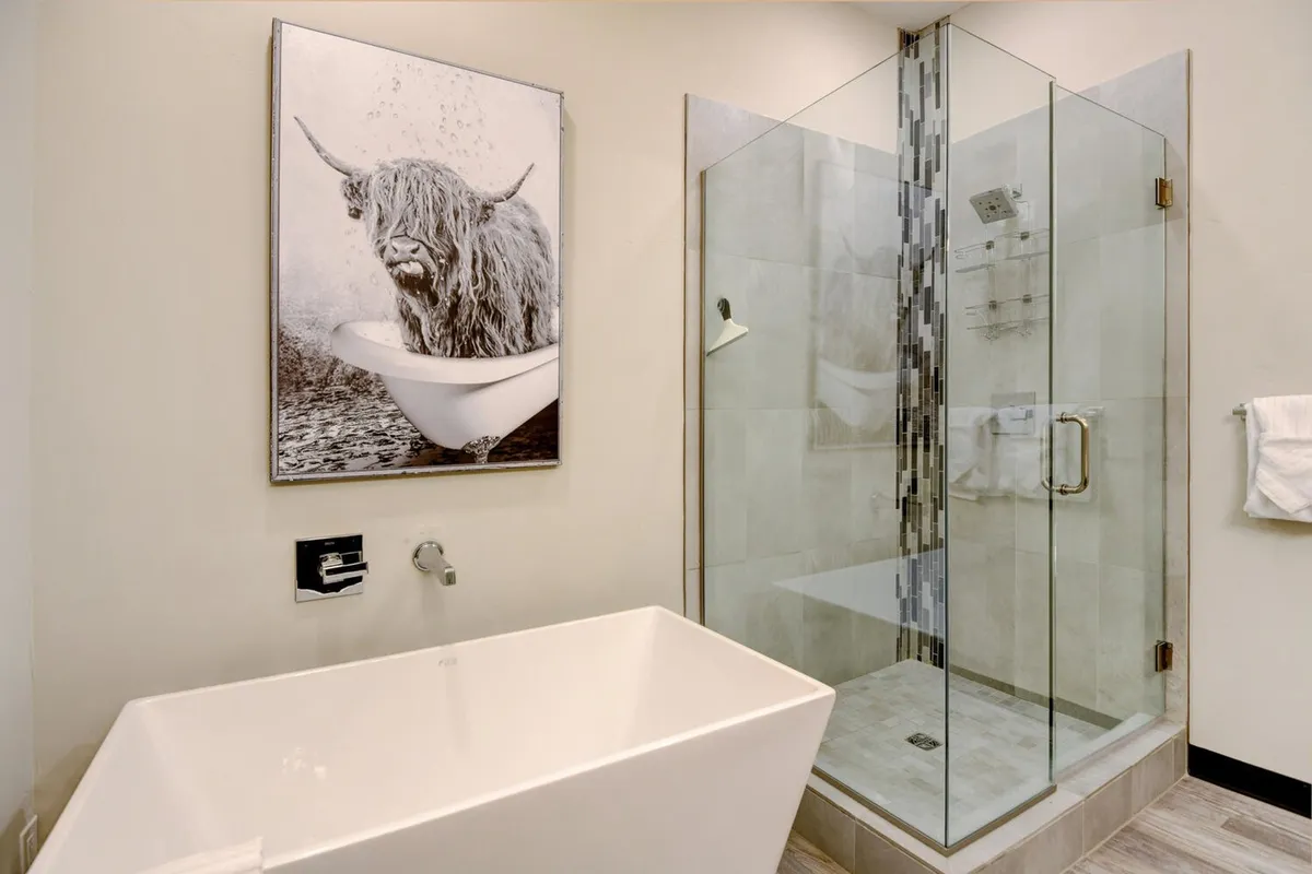 A contemporary bathroom featuring a freestanding bathtub, a sleek glass shower enclosure, and a distinctive artwork of a highland cow. The space radiates tranquility and luxury, ideal for relaxing after a day of adventure.