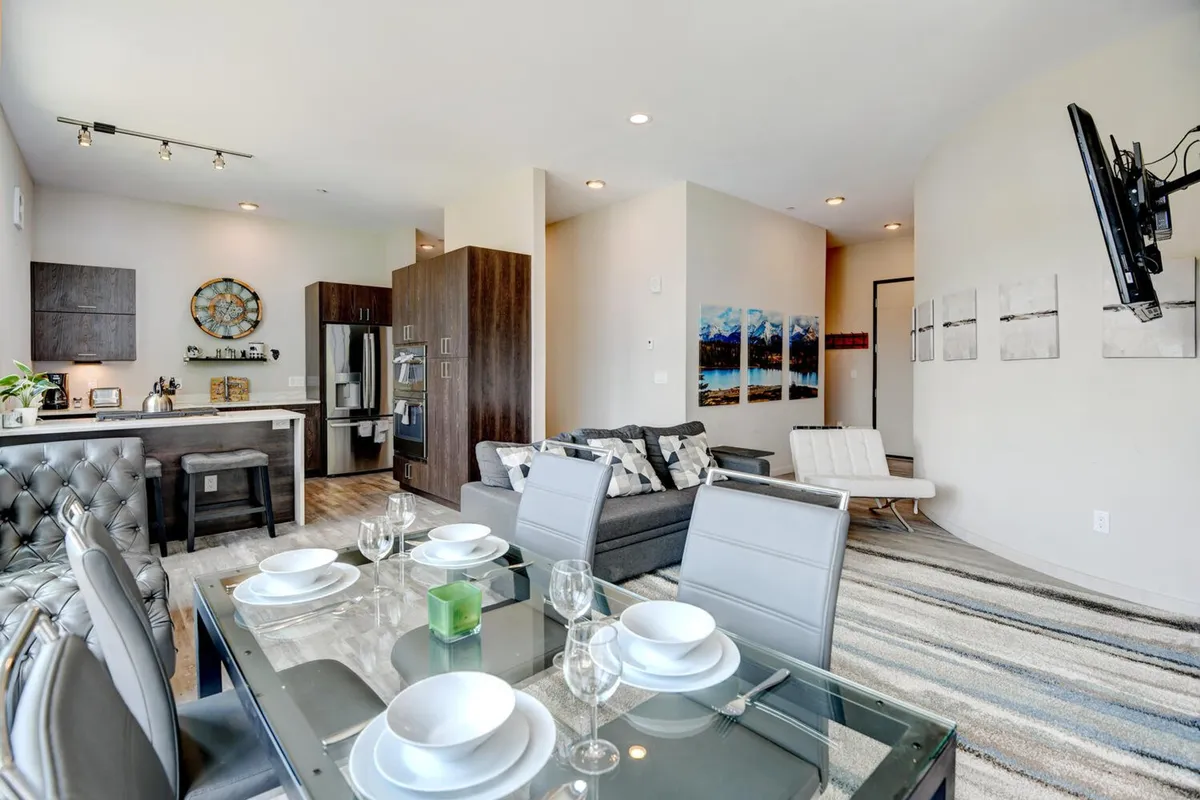 This interior image presents a stylish open-concept area that combines a dining table set for a meal, modern kitchen appliances, and a comfortable seating area. The arrangement invites social gatherings, and the soft natural light streaming through large windows suggests a warm, welcoming atmosphere with a contemporary touch.