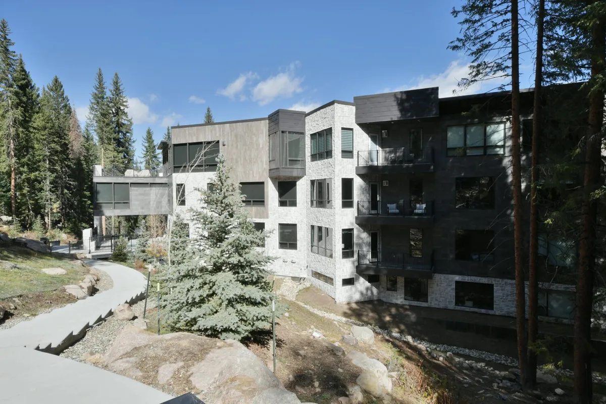 This exterior image showcases a modern building surrounded by lush trees, with a pathway leading up to the entrance. The scene evokes the natural beauty and tranquility of the area, while the architectural style hints at both comfort and adventure. The setting suggests easy access to nearby trails and outdoor activities.