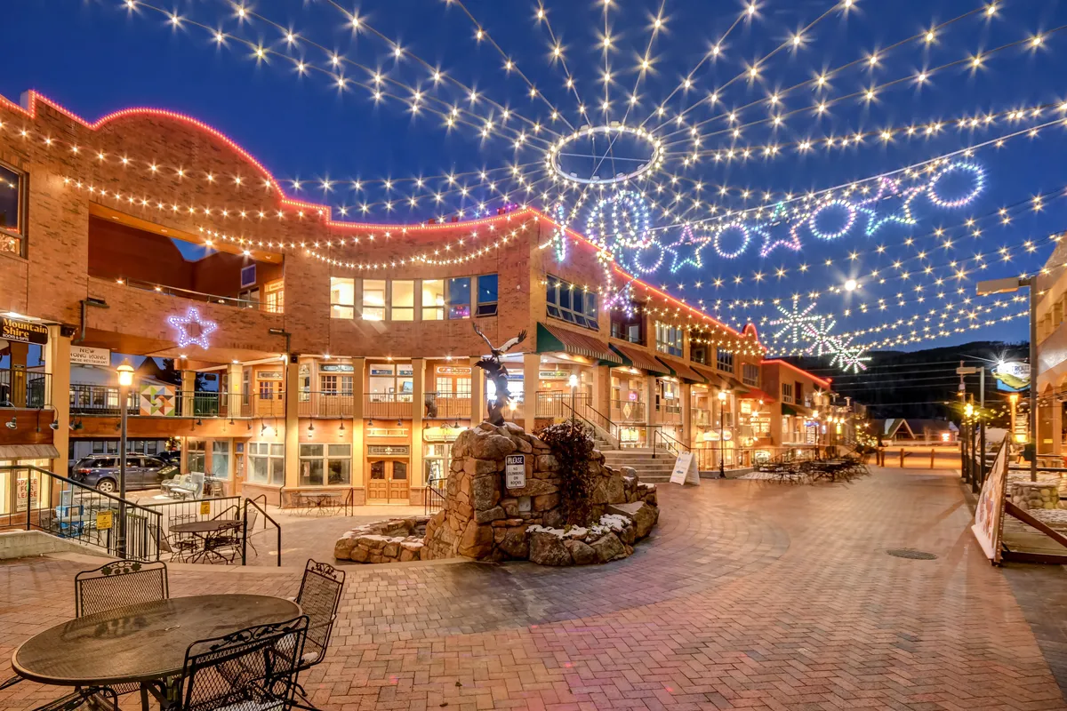 An inviting outdoor plaza is lit up with string lights and decorative stars, featuring a stone fountain and ample seating. The area feels festive and lively, suggesting social gatherings, events, and community celebrations.