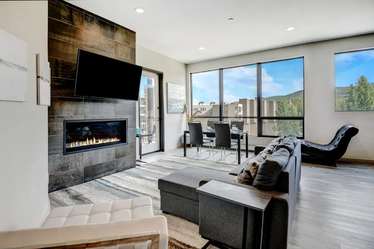 The hero image showcases a chic, contemporary living space featuring a modern fireplace, expansive windows framing breathtaking mountain sunset views, plush seating, and an inviting dining area. The scene evokes warmth and sociability, ideal for relaxing by the fire, gathering with loved ones, or savoring a meal with a stunning backdrop.