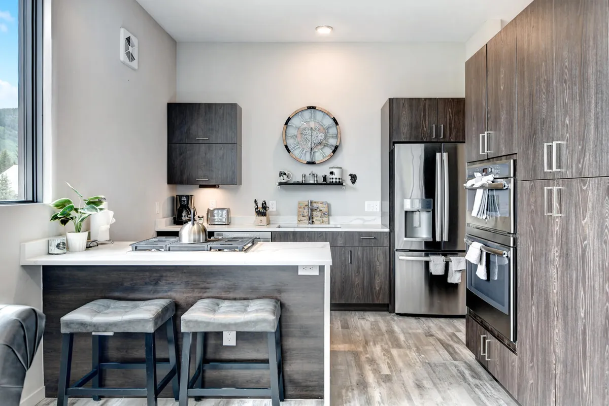 A bright, inviting kitchen space featuring a countertop with bar stools, modern kitchen appliances, a wall clock, and open shelves decorated tastefully. The setup is ideal for family meal preparations and gatherings, with plenty of space for cooking and social activities.