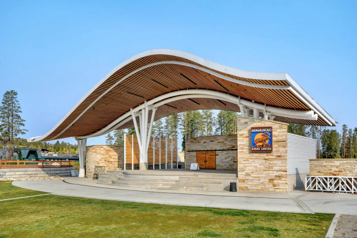 An exterior scene featuring a striking curved-roof structure with stone walls and wooden slats, set amid the natural landscape. The area is designed for concerts, community events, and gatherings, further reinforcing the theme of year-round activities.