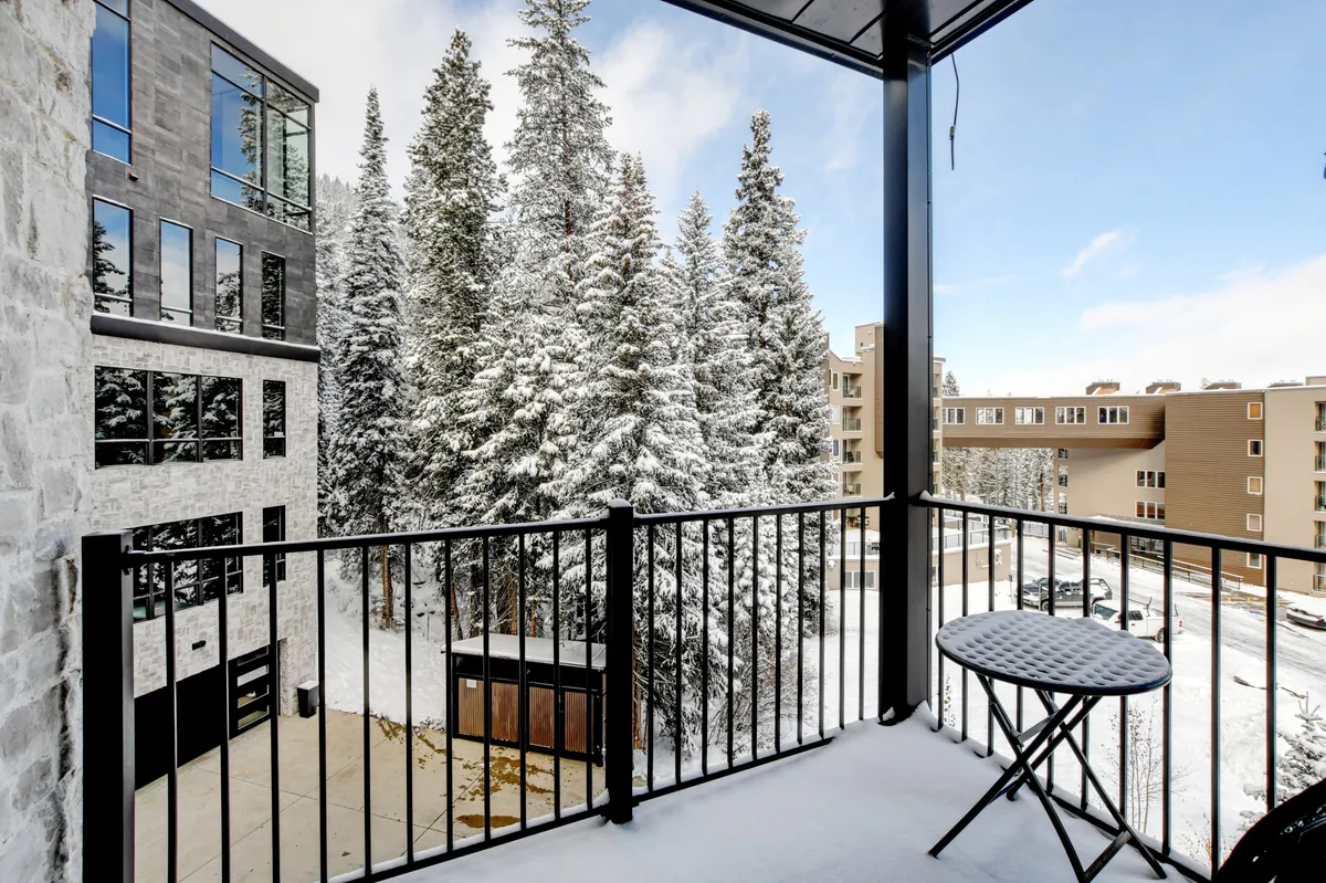 A cozy exterior balcony framed by a railing, overlooking snow-covered trees. The small table and chair invite quiet mornings or evenings spent enjoying the tranquil winter scenery. The scene captures the allure of private outdoor space with picturesque mountain surroundings.