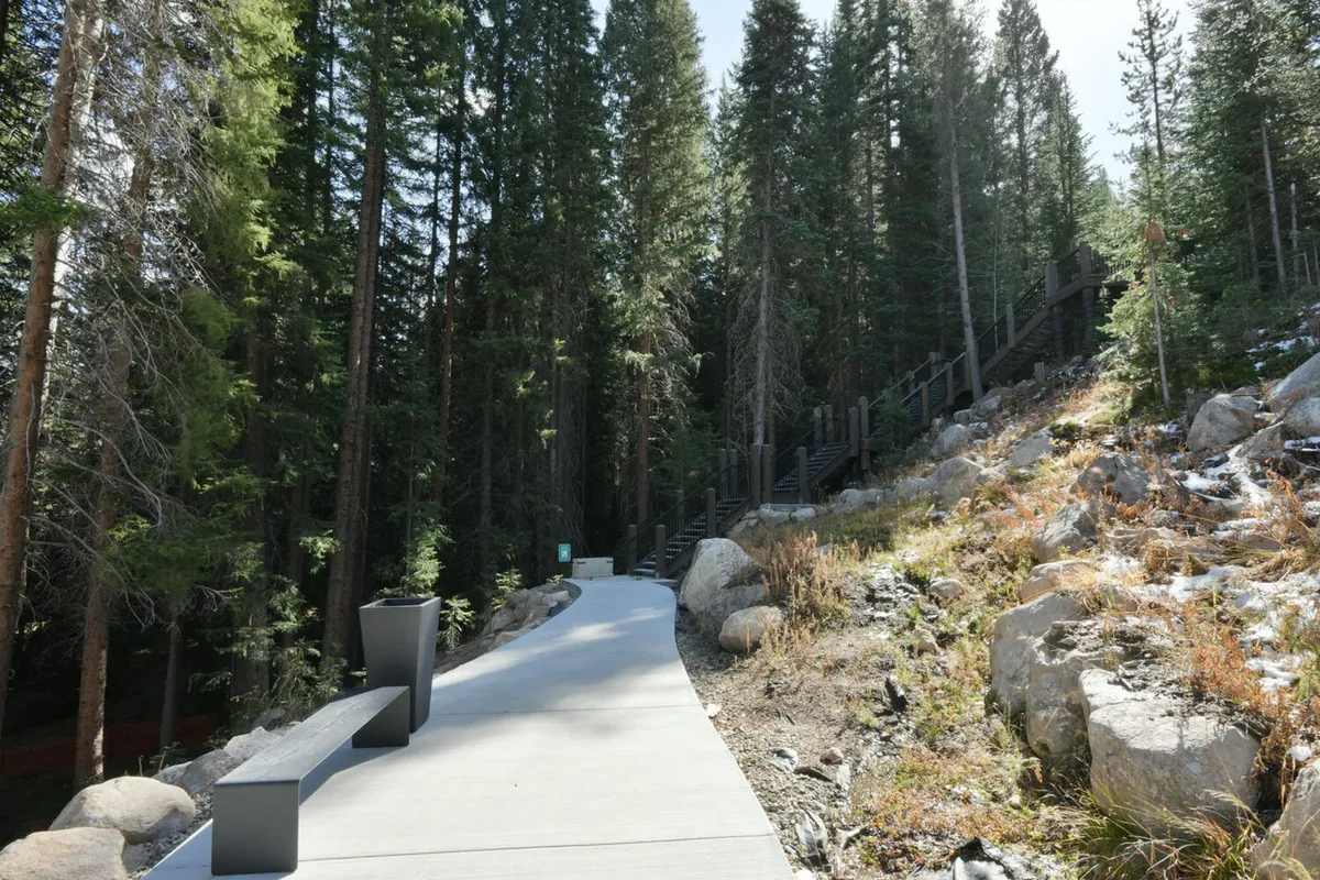 A peaceful exterior setting with a winding concrete pathway surrounded by lush forest and natural rock formations. Wooden stairs invite exploration, and the area exudes a sense of immersion in nature.