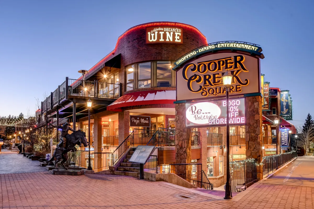 A vibrant streetscape at the resort base featuring the Cooper Creek Square sign, outdoor dining, and lively architecture. The scene captures the heart of the local dining, shopping, and entertainment options available just steps away from the retreat.